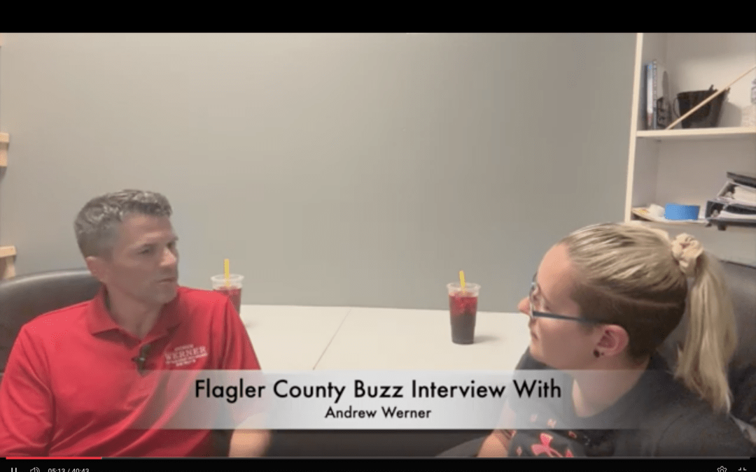 Two people seated at a table in Palm Coast, engaged in an interview The man on the left wears a red shirt, while a woman with glasses sits on the right Two drinks with straws are on the table, and a text overlay reads "Flagler County Buzz Interview With Andrew Werner